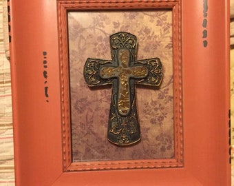 May include: A framed decorative cross. The frame is a distressed, muted orange-red color. The cross is metallic with ornate detailing and a gold-colored center. The background is a floral pattern in muted tones.