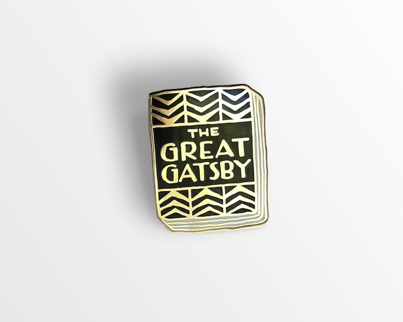K&ouml;nnte beinhalten: Eine goldfarbene Emaille-Anstecknadel in Form eines Buches mit schwarzem Einband und goldener Aufschrift "THE GREAT GATSBY". Der Einband hat ein Muster aus goldenen Chevron.