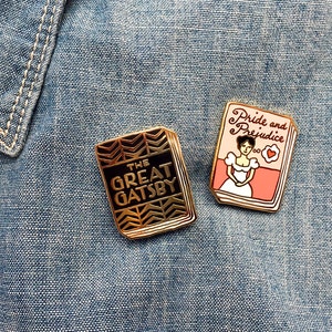 K&ouml;nnte beinhalten: Zwei goldfarbene Emaille-Pins mit klassischen Buchumschl&auml;gen. Der erste Pin zeigt ein schwarzes und goldenes Design mit dem Text "The Great Gatsby". Der zweite Pin zeigt ein rosa und wei&szlig;es Design mit dem Text "Pride and Prejudice".