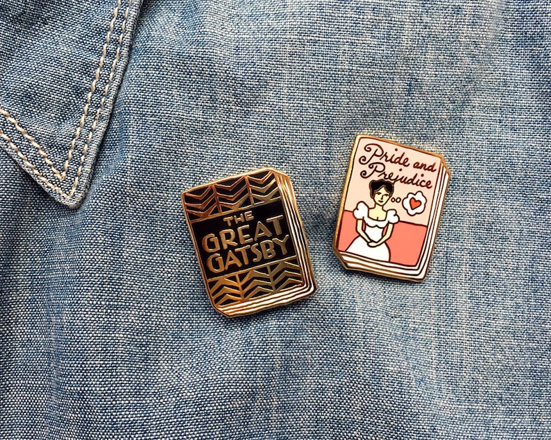 K&ouml;nnte beinhalten: Zwei goldfarbene Emaille-Pins mit klassischen Buchumschl&auml;gen. Der erste Pin zeigt ein schwarzes und goldenes Design mit dem Text "The Great Gatsby". Der zweite Pin zeigt ein rosa und wei&szlig;es Design mit dem Text "Pride and Prejudice".