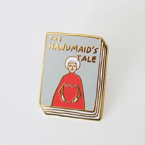 May include: Enamel pin featuring the cover of "The Handmaid's Tale." The pin depicts a woman in a red cloak and white bonnet, with the book title in gold lettering on a gray background. The pin has a gold border.
