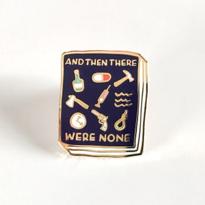 以下が含まれることがあります： 金色の縁取りが施された濃い青色のエナメルピン。小さな金色のアイコンと「AND THEN THERE WERE NONE」の文字が特徴です。アイコンには、ボトル、ピル、ハンマー、斧、注射器、波、時計、銃、そして縄が含まれています。