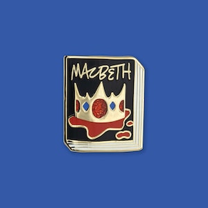 May include: A rectangular enamel pin shaped like a book with a black cover, gold edges, and the word Macbeth written in gold script. The cover features a gold crown with red glitter accents resting on a red blood-like splatter design.