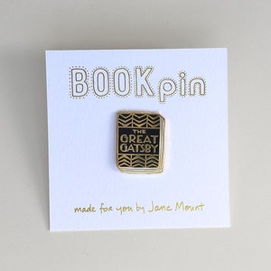 K&ouml;nnte beinhalten: Goldene Anstecknadel mit einem schwarzen Buch mit dem Text "The Great Gatsby" auf dem Cover. Die Anstecknadel befindet sich auf einer wei&szlig;en Karte mit dem Text "BOOK pin" und "made for you by Jane Mount".