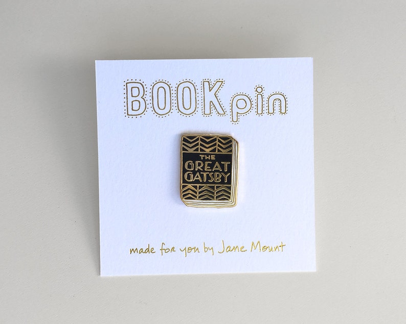 K&ouml;nnte beinhalten: Goldene Anstecknadel mit einem schwarzen Buch mit dem Text "The Great Gatsby" auf dem Cover. Die Anstecknadel befindet sich auf einer wei&szlig;en Karte mit dem Text "BOOK pin" und "made for you by Jane Mount".