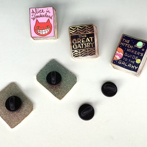 K&ouml;nnte beinhalten: Satz von f&uuml;nf Emaille-Pins mit Buchumschl&auml;gen. Die Pins sind goldfarben mit bunten Emaille-Details. Die Buchtitel lauten "Alice im Wunderland", "Der gro&szlig;e Gatsby" und "Per Anhalter durch die Galaxis".
