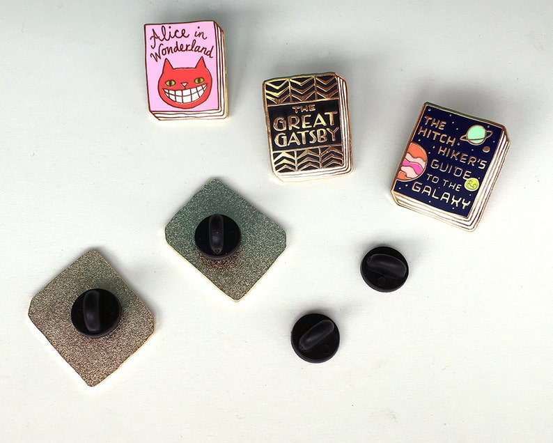 K&ouml;nnte beinhalten: Satz von f&uuml;nf Emaille-Pins mit Buchumschl&auml;gen. Die Pins sind goldfarben mit bunten Emaille-Details. Die Buchtitel lauten "Alice im Wunderland", "Der gro&szlig;e Gatsby" und "Per Anhalter durch die Galaxis".