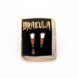 May include: A black enamel pin shaped like a book with gold trim. The book cover says "DRACULA" in gold lettering. There are two red and white fangs with a red blood drop on the cover.