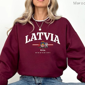 May include: Maroon sweatshirt featuring the word "LATVIA" in white. Below the word is a graphic of the Latvian coat of arms and the city name "RIGA" with coordinates. The sweatshirt has long sleeves and a crew neck.