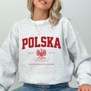 Könnte beinhalten: Ein hellgraues Sweatshirt mit dem Wort "POLSKA" in fetten roten Buchstaben. Darunter befindet sich ein rotes polnisches Adleremblem mit dem Text "EST. 1918" und "WARSZAWA RZECZPOSPOLITA POLSKA". Das Sweatshirt wird mit einer blauen Jeans getragen.
