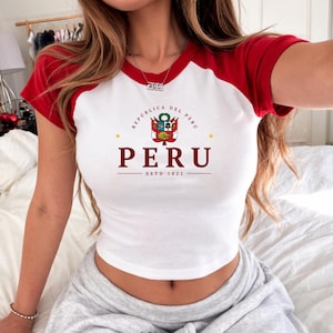 May include: White crop top with red short sleeves featuring the text "PERU" and the Peruvian coat of arms. The shirt also includes the text "REPUBLICA DEL PERU" and "ESTD 1821".
