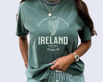 May include: A sage green short-sleeve t-shirt featuring a white graphic of a harp and the text Republic of Ireland, Ireland, Estd 1937, Dublin. The shirt is paired with green and white gingham patterned shorts. The text Blue Spruce is visible.