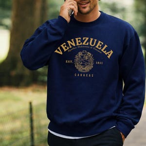May include: Navy blue sweatshirt with gold lettering and a crest design. The word "VENEZUELA" arches across the top, with "Rep&uacute;blica Bolivariana de Venezuela" above the crest. Below the crest are the words "EST. 1811 CARACAS".