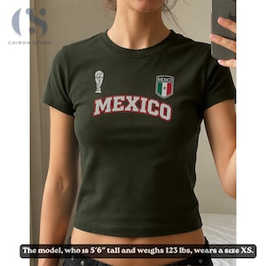 Op de afbeelding: Een donker olijfgroen T-shirt met korte mouwen en het woord MEXICO in witte letters met een rode omlijning. Boven de tekst staat een wit trofee-icoon en een schildvormig embleem in de kleuren van de Mexicaanse vlag. Model is 168 cm en 56 kg.