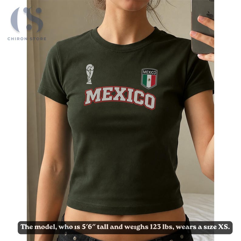 Op de afbeelding: Een donker olijfgroen T-shirt met korte mouwen en het woord MEXICO in witte letters met een rode omlijning. Boven de tekst staat een wit trofee-icoon en een schildvormig embleem in de kleuren van de Mexicaanse vlag. Model is 168 cm en 56 kg.
