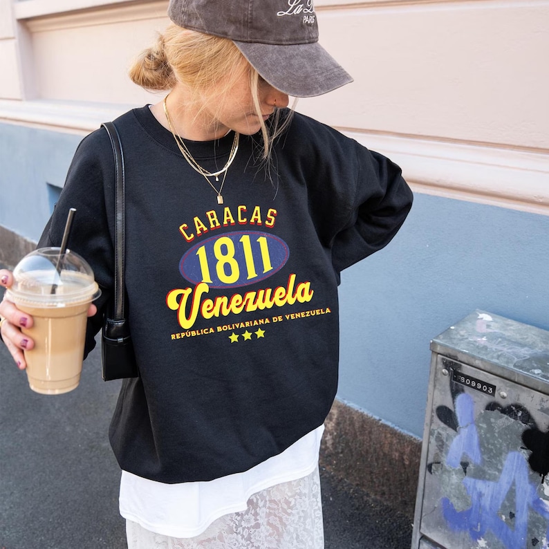May include: Black sweatshirt with the text "CARACAS 1811 Venezuela" in yellow and blue. The sweatshirt is worn with a gray cap and a white skirt. A person is holding a beverage in a clear cup.