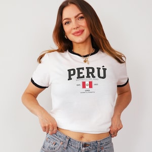 Peut inclure: T-shirt blanc à col rond et bordures noires avec le mot "PERÚ" en gras. Le drapeau péruvien et le texte "EST. 1821 LIMA" sont en dessous. Le t-shirt est porté par une personne.