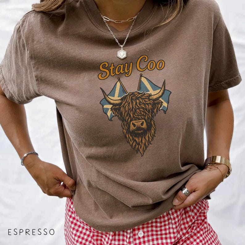 K&ouml;nnte beinhalten: Braunes T-Shirt mit dem Schriftzug "Stay Coo" &uuml;ber einer Grafik einer schottischen Hochlandkuh und schwedischen Flaggen. Das Shirt hat einen Rundhalsausschnitt und kurze &Auml;rmel. Unten ist das Wort "Espresso" zu sehen.