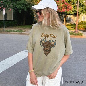 K&ouml;nnte beinhalten: Khakifarbenes T-Shirt mit dem Text "Stay Coo" &uuml;ber einer braunen Highland-Kuh-Grafik mit zwei schottischen Flaggen. Das Shirt hat eine lockere Passform und kurze &Auml;rmel. Der Text "KHAKI GREEN" befindet sich unten rechts im Bild.