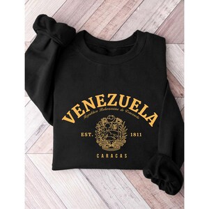 May include: Black sweatshirt with the word "VENEZUELA" in gold lettering across the chest. Below the word is a gold emblem and the words "Republica Bolivariana de Venezuela", "EST. 1811", and "CARACAS".