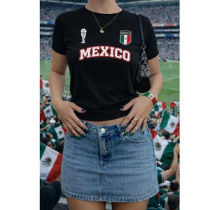 Op de afbeelding: Een zwart T-shirt met korte mouwen met het woord MEXICO in witte letters met een rode rand, een klein wit trofee-icoon en een Mexicaans vlag-embleem. Het shirt wordt gecombineerd met een blauwe spijkerrok en een zwarte schoudertas met logopatroon.