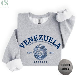 May include: A light gray sweatshirt with the word "VENEZUELA" in navy blue arched across the top. Below the word is a navy blue coat of arms and the words "EST. 1811 CARACAS". The sweatshirt has white fleece lining the inside and the sleeves are folded up.