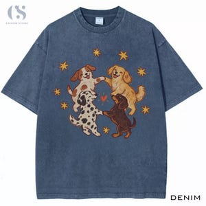 Puede incluir: Una camiseta azul con efecto lavado al ácido que presenta un gráfico de cuatro perros tomados de las patas en círculo, rodeados de estrellas amarillas y un pequeño corazón rojo. Los perros incluyen uno manchado marrón y blanco, uno dorado, un dálmata y uno marrón.