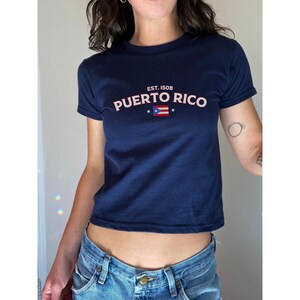 以下が含まれることがあります： 白と赤のプエルトリコの国旗のグラフィックと、ピンク色の「EST. 1508 PUERTO RICO」の文字が入った、ネイビーブルーのクロップドTシャツです。