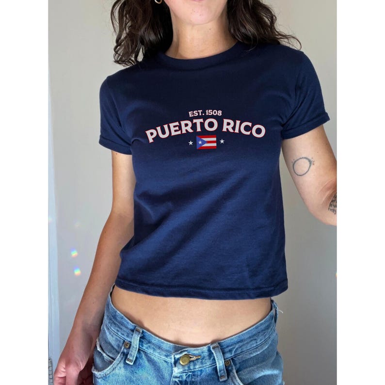 以下が含まれることがあります： 白と赤のプエルトリコの国旗のグラフィックと、ピンク色の「EST. 1508 PUERTO RICO」の文字が入った、ネイビーブルーのクロップドTシャツです。