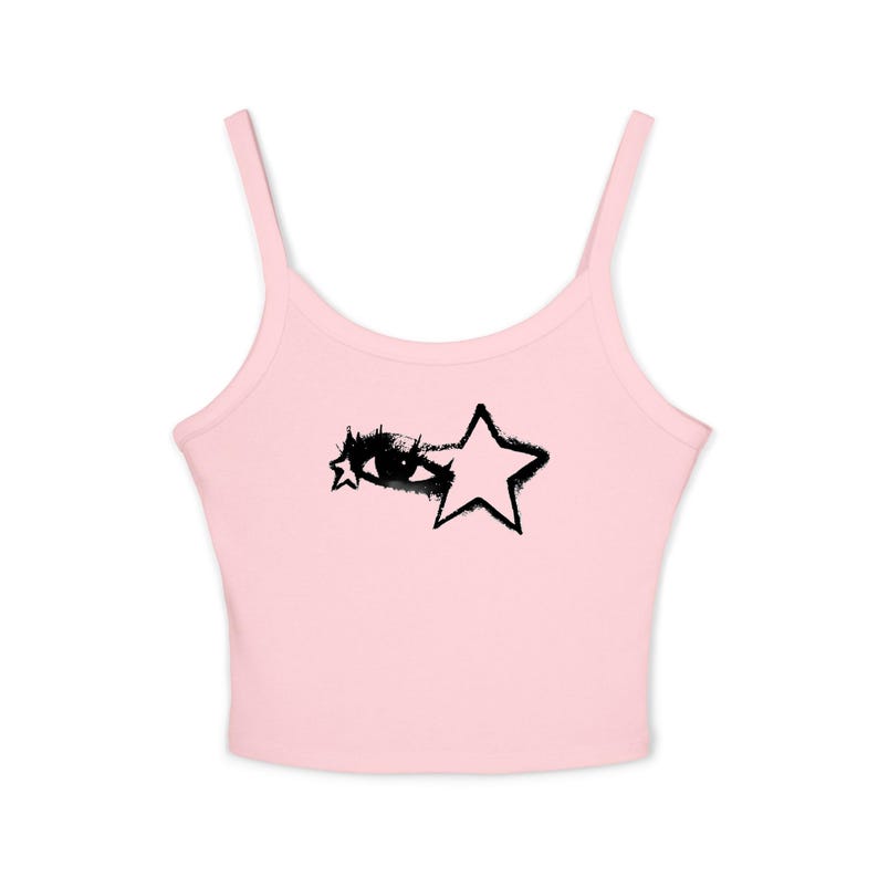 May include: A light pink camisole top with thin straps. The top features a black graphic of an eye and a star. The design is centered on the front of the top. The top is made of a soft, ribbed material.