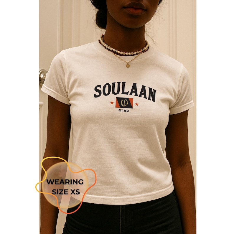 May include: White t-shirt with the word "SOULAAN" in black, a flag graphic, and "EST. 1865" in red. The shirt has short sleeves and is worn with a pearl necklace. The person is wearing size XS.