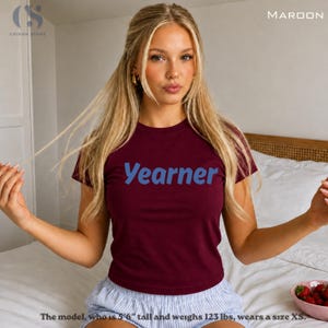 Può includere: Una maglietta bordeaux con la scritta "Yearner" in blu. La modella indossa una taglia XS. La maglietta è a tinta unita e ha un girocollo. La modella è seduta su un letto.