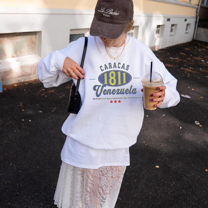May include: A white sweatshirt with the text "CARACAS 1811 Venezuela" in blue and yellow. The person is wearing a brown baseball cap with "La Po&eacute;sie Paris" printed on it. The person is also wearing a white skirt and holding a black purse and a coffee drink.
