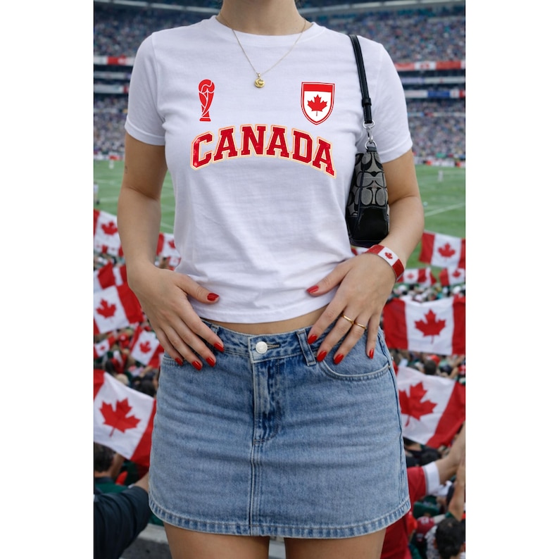 Peut inclure: T-shirt blanc avec le mot "CANADA" en rouge, un embl&egrave;me de feuille d'&eacute;rable rouge et un graphisme de troph&eacute;e de football. La tenue comprend une jupe en jean. Un sac &agrave; main noir est port&eacute; &agrave; l'&eacute;paule.