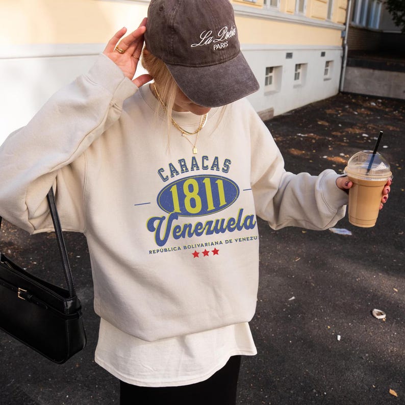 May include: A beige sweatshirt with the text "CARACAS 1811 Venezuela" in blue and yellow. The sweatshirt is paired with a brown cap that says "La Paris". A black purse and a drink in a clear cup are also visible.