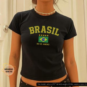 May include: Black crop top with the word "BRASIL" in yellow block letters, a Brazilian flag graphic, and the text "RIO DE JANEIRO". The shirt has a crew neck and short sleeves. The model is wearing size XS.