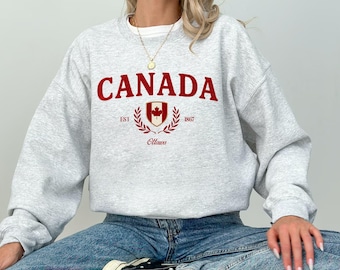Pode incluir: Um moletom cinza claro com a palavra "CANADA" em letras vermelhas. Abaixo, um brasão vermelho com uma folha de bordo e as palavras "EST 1867 Ottawa". O moletom é feito de um material macio.