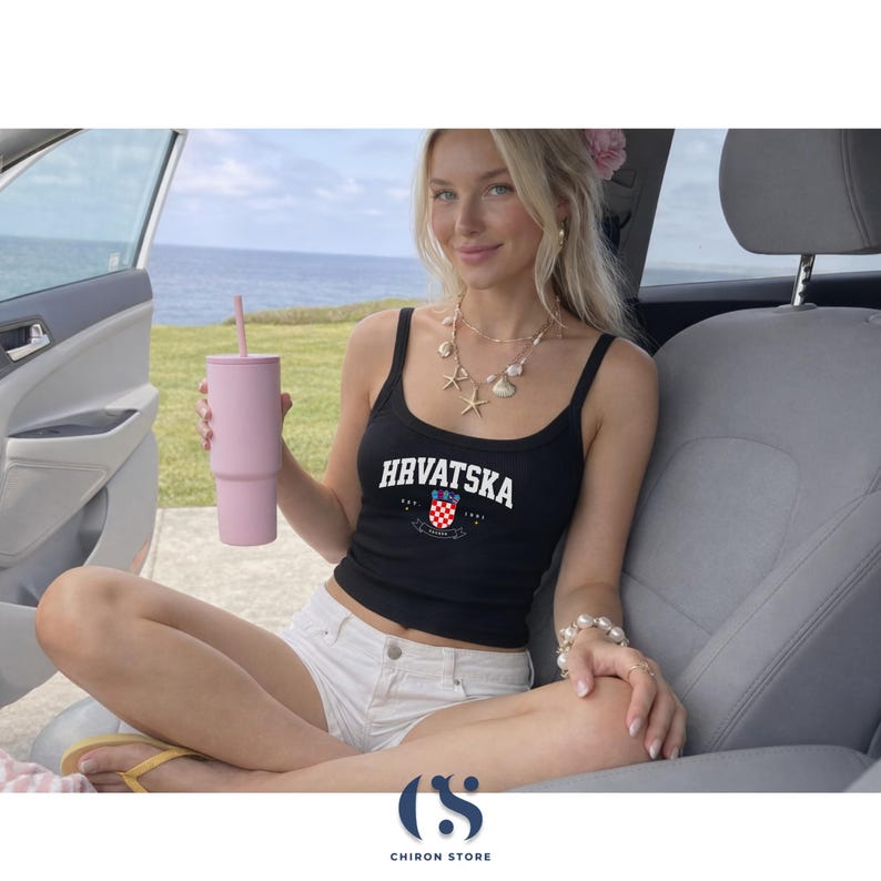 Op de afbeelding: Zwarte tanktop met het woord "Hrvatska" in het wit, een Kroatisch wapenschild en een roze beker met een rietje. De persoon draagt een witte short en een schelpenketting.