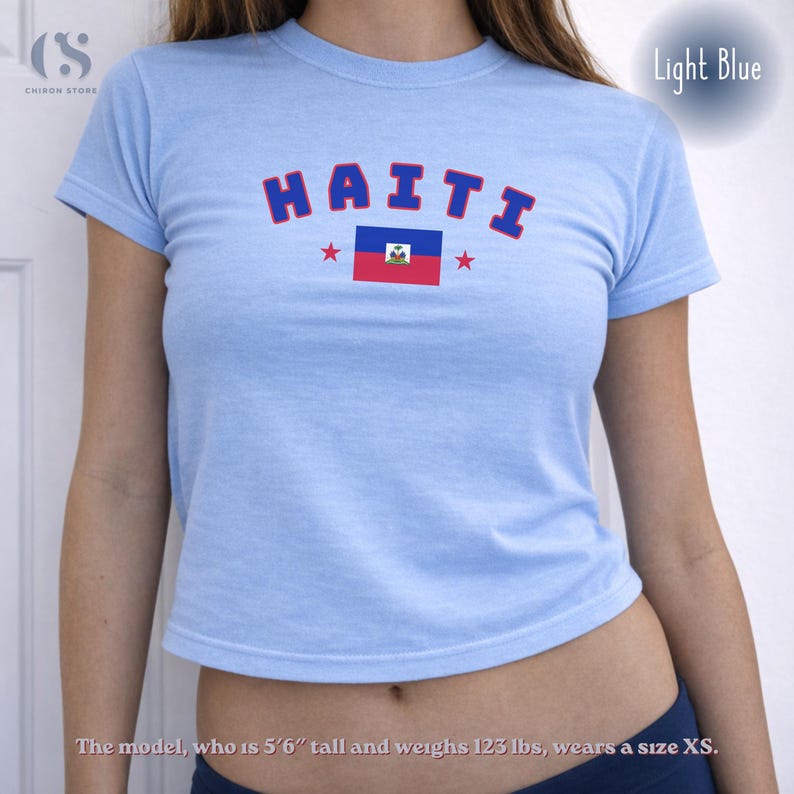 May include: Light blue crop top with the word "HAITI" in navy blue arched letters with red stars. The flag of Haiti is centered below the word. The shirt is a light blue color.