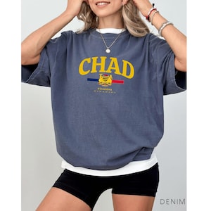May include: A denim blue t-shirt with the word "CHAD" in yellow block letters. Below the word is a crest design with the text "N'DJAMENA". The shirt has short sleeves and a white undershirt is visible.
