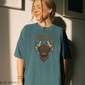 K&ouml;nnte beinhalten: Blaugr&uuml;nes T-Shirt mit einem Highland-Kuh-Motiv und dem Text "Stay Coo" in goldfarbenen Buchstaben. Das Design beinhaltet zwei schottische Flaggen. Das Shirt hat eine lockere Passform.