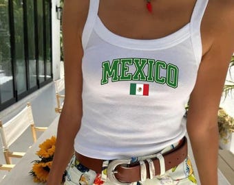 May include: White vest top featuring the word "MEXICO" in green lettering and a small Mexican flag. The top has a scoop neckline and thin straps. The model is wearing a brown belt with a silver buckle.