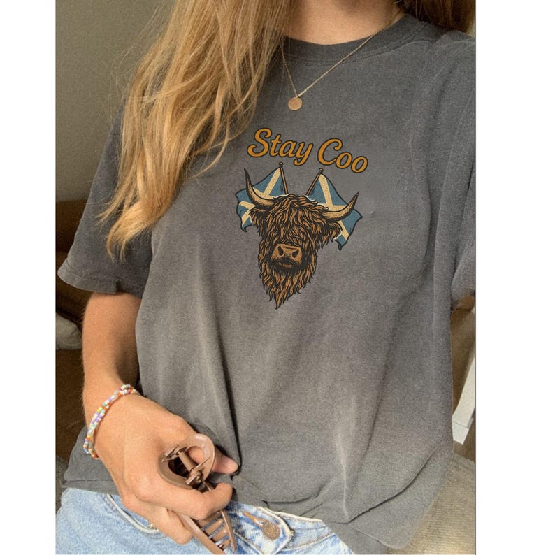 K&ouml;nnte beinhalten: Ein graues T-Shirt mit einem Highland-Kuh-Motiv und dem Text "Stay Coo". Das Design beinhaltet die schottische Flagge. Dies ist ein Freizeitkleidungsst&uuml;ck.