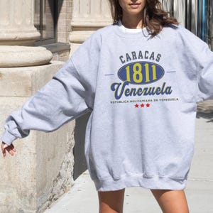 May include: A gray sweatshirt with the text "CARACAS 1811 Venezuela" in blue and yellow. The sweatshirt also has the text "REPUBLICA BOLIVARIANA DE VENEZUELA" and three red stars. The sweatshirt is a casual style.