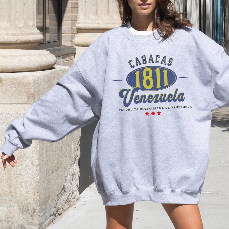 May include: A gray sweatshirt with the text "CARACAS 1811 Venezuela" in blue and yellow. The sweatshirt also has the text "REPUBLICA BOLIVARIANA DE VENEZUELA" and three red stars. The sweatshirt is a casual style.