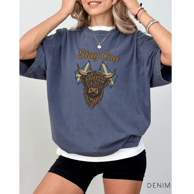 K&ouml;nnte beinhalten: Denimblaues T-Shirt mit dem Text "Stay Coo" &uuml;ber einer Highland-Kuh-Grafik und zwei schottischen Flaggen. Das Shirt hat eine lockere Passform, und ein wei&szlig;es Unterhemd ist am Ausschnitt und am Saum sichtbar.