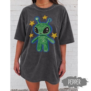 May include: A charcoal grey short-sleeve t-shirt featuring a central graphic of a green alien character with large black eyes. The alien is surrounded by four yellow stars, and the entire design has a textured, crayon-like appearance with blue outlines.