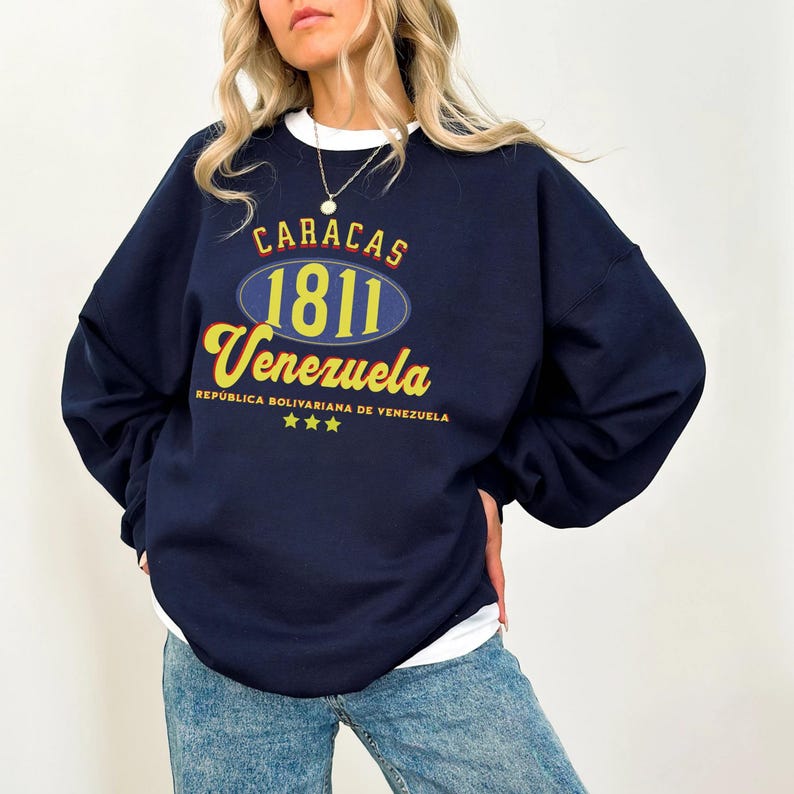May include: Navy blue sweatshirt with the text "CARACAS 1811 Venezuela" in yellow and red lettering. The sweatshirt has a white collar and a design that includes three yellow stars and the text "REPUBLICA BOLIVARIANA DE VENEZUELA".