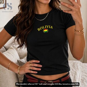 May include: Black t-shirt with the word "BOLIVIA" in gold lettering above a Bolivian flag emblem. The shirt is short-sleeved and has a crew neck. The model is wearing the shirt. The text at the bottom says the model is 5'6" tall and weighs 123 lbs, wearing a size XS.