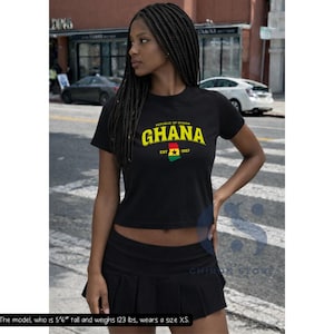 May include: A black short-sleeved crop top featuring the text Republic of Ghana GHANA EST 1957 in yellow and white, with a graphic of the Ghana flag in the center. The shirt is paired with a black pleated mini skirt.